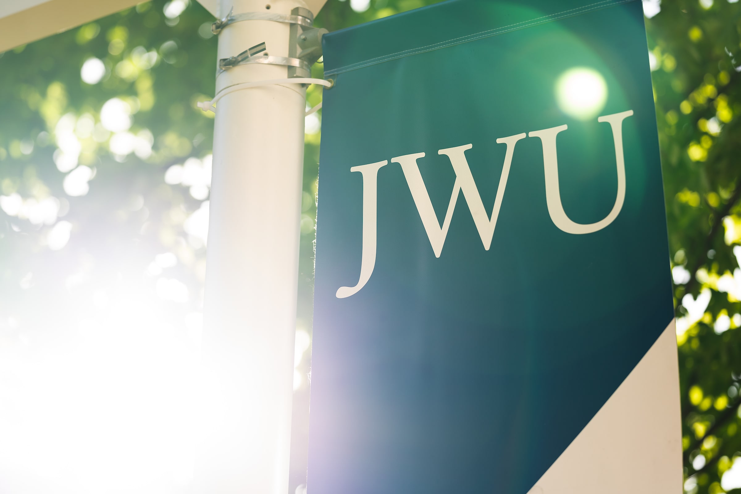 JWU blue and white shield logo on banner next to a tree with green leaves