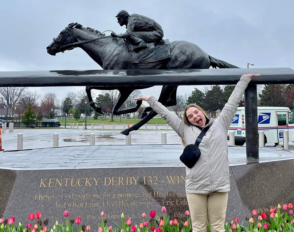 Female student raises her arms in front of Kentucky Derby statue.