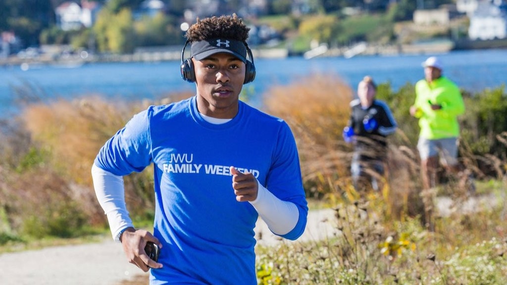 JWU student in blue t-shirt running on an outdoor path with Narragansett Bay in the background.