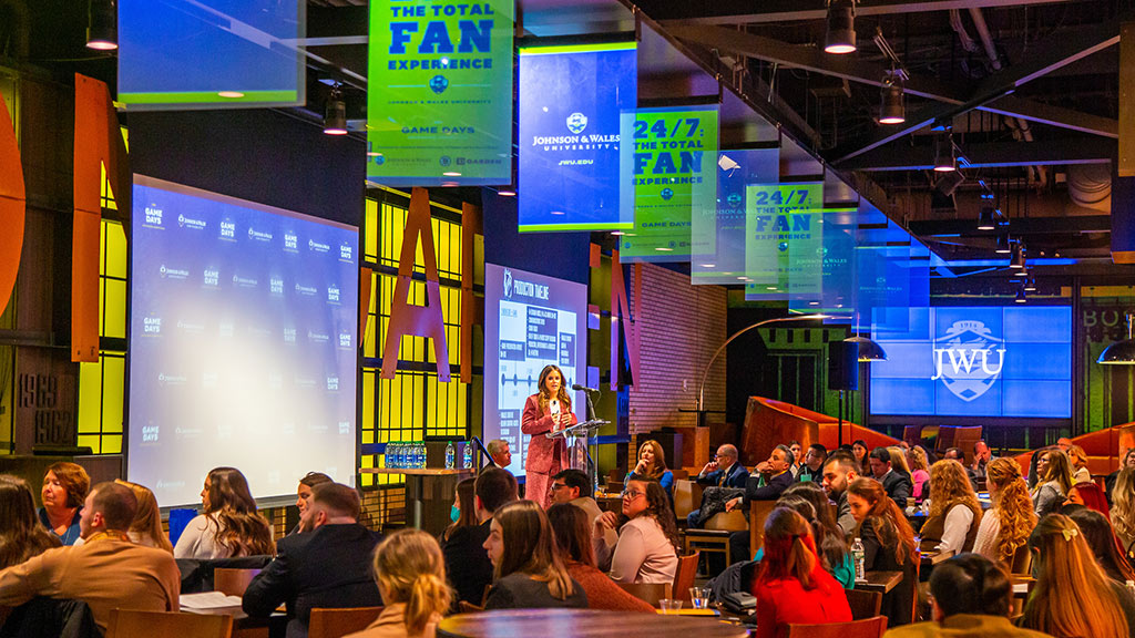 Renee Riva speaking to the crowd at TD Garden Legends restaurant, surrounded by JWU Game Days signage