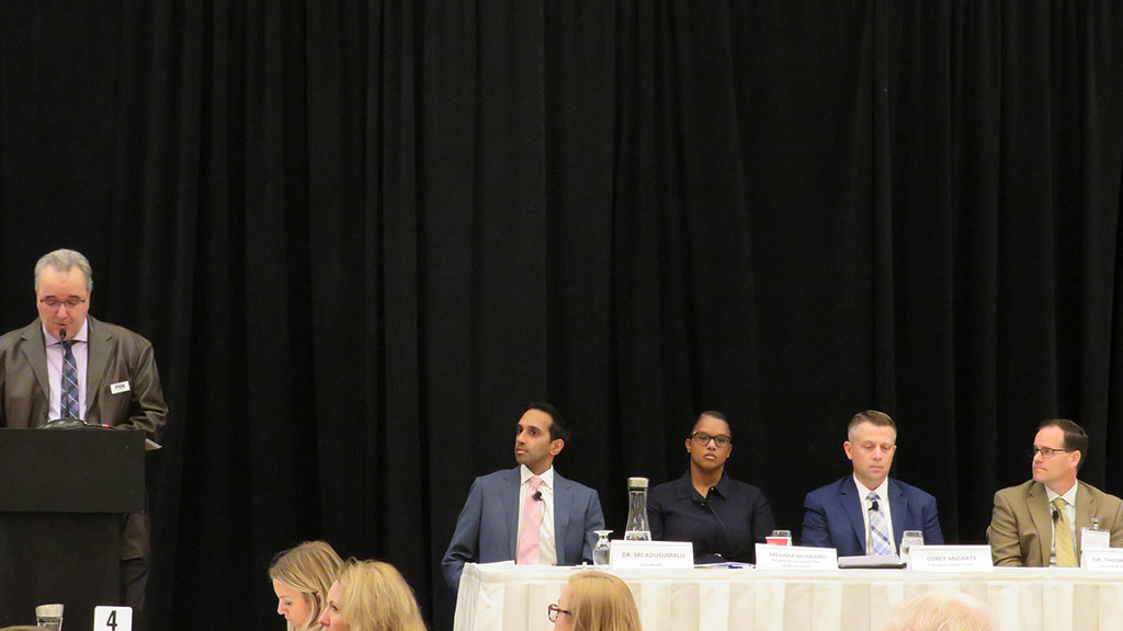 photo of the PBN moderator reading at a podium while four panelists, including Thomas Meehan on the right, sit at a table on stage