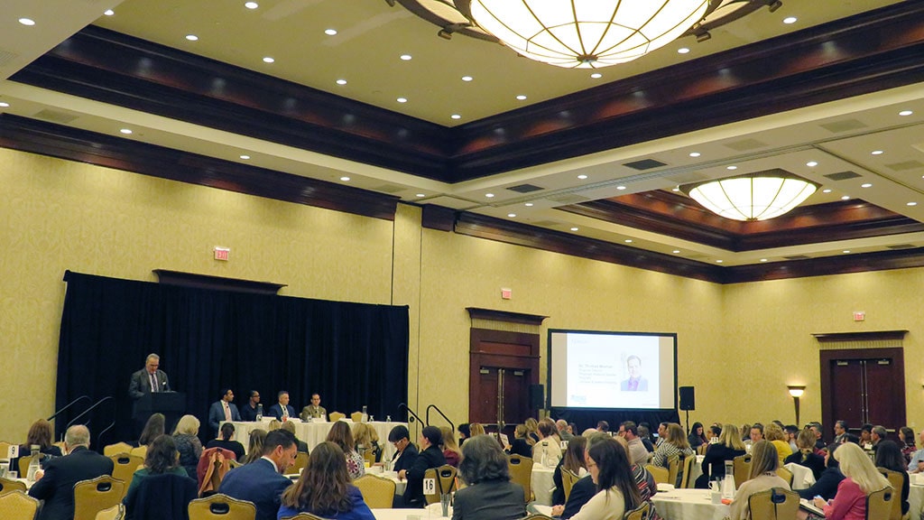 a view of the healthcare summit with dozens of people sitting at tables while the panelists are on a table on stage