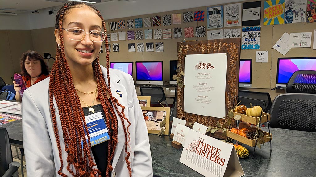 Erika Morais '23 poses with some of the elements her team graphic designed for a culinary students' "three sisters" dining experience