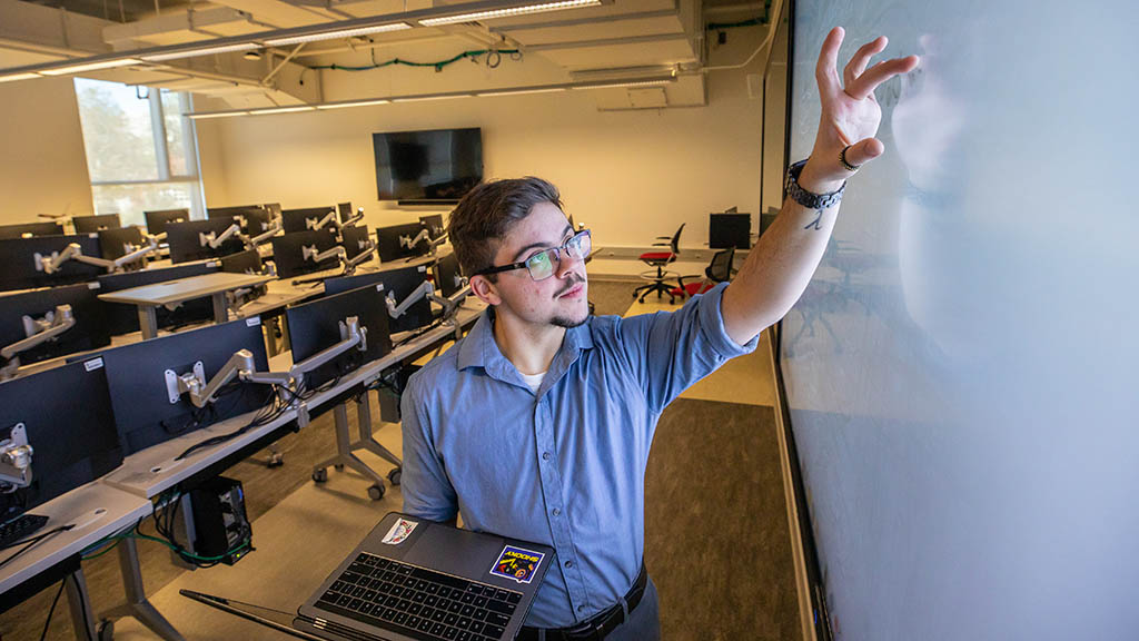 a closeup photo of Jae Grenier '23 holding his laptop while writing on a large screen in the computer science lab