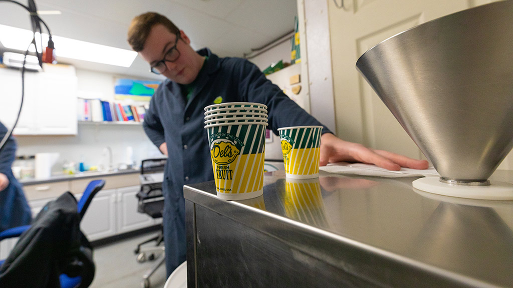 photo of a metal machine in the forefront topped with several Del’s lemonade cups, with Caleb Atkinson ’24 in the background