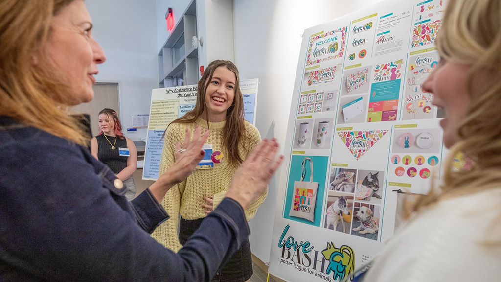 Julia Shiels standing in front of a board with her design work and talking with two women.