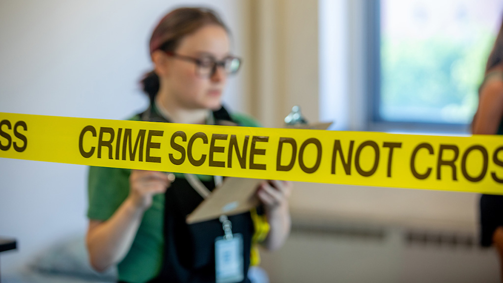 Students investigate a mock crime scene, yellow tape with the words "Crime scene do not cross"covers some of the image