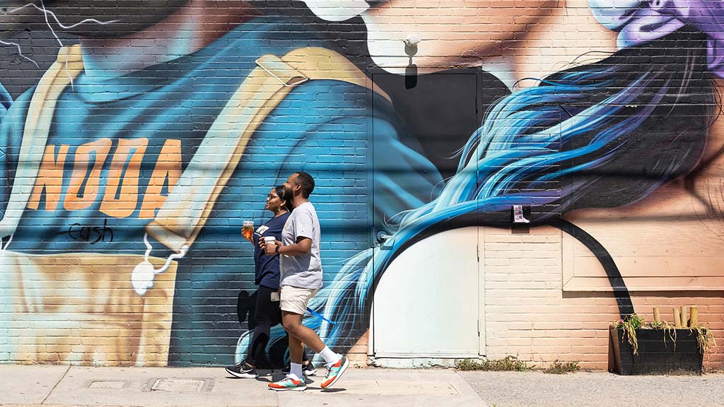 JWU students stroll while drinking iced coffees in front of a vibrant mural.