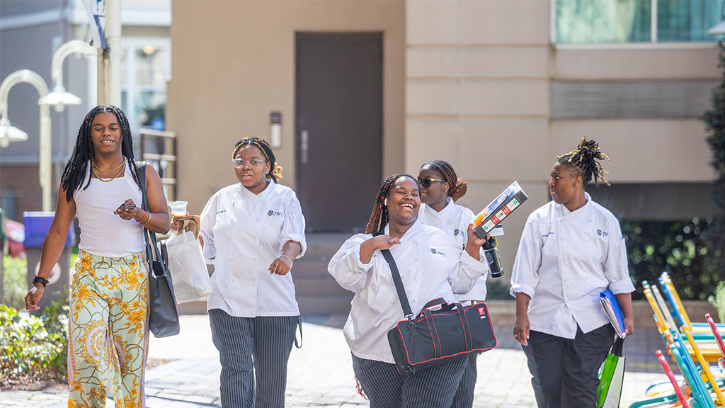 Students in chef uniforms, who are milling and carrying bags walking through campus on a sunny day.