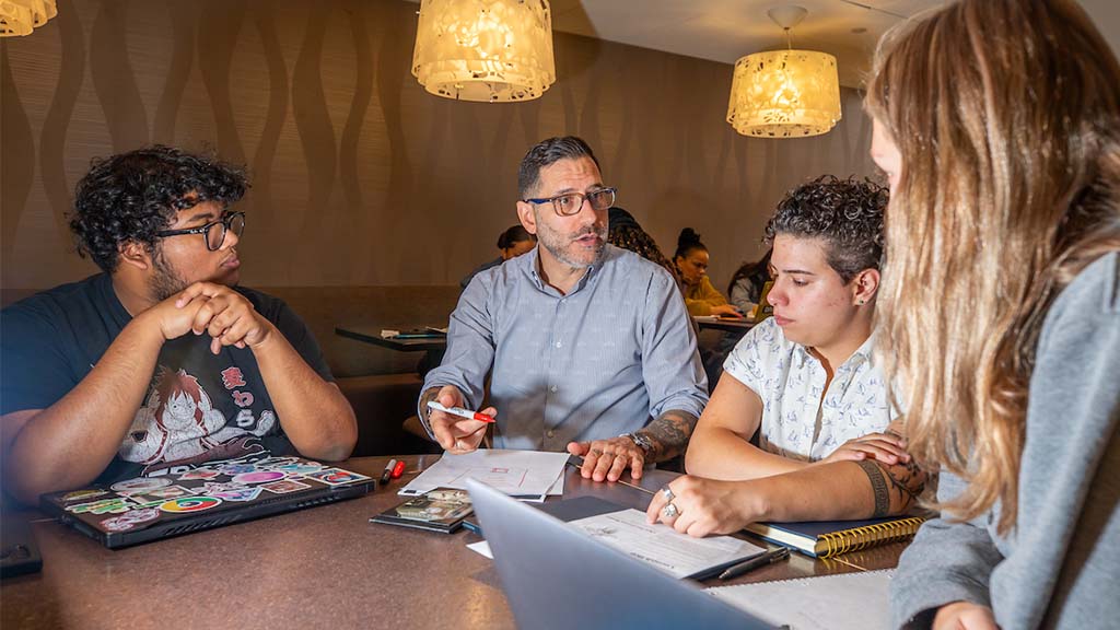 A JWU faculty member sits with three students at a table in Bistro 61, guiding them through notes and assignments during a collaborative study session.
