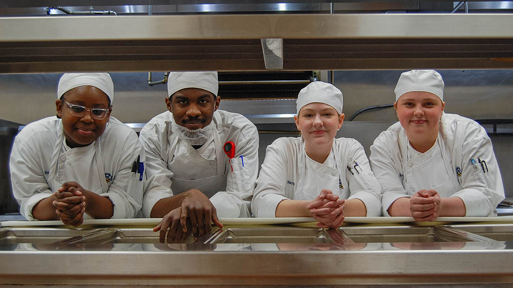 Students in chef whites in a culinary lab smile.