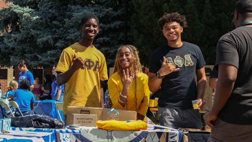 Three students, one sitting and two standing, with Greek Life shirts on smiling