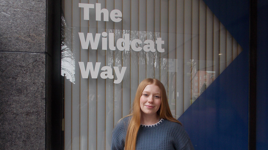 Katie McPartlin '28 stands under the words "The Wildcat Way" on a JWU wall