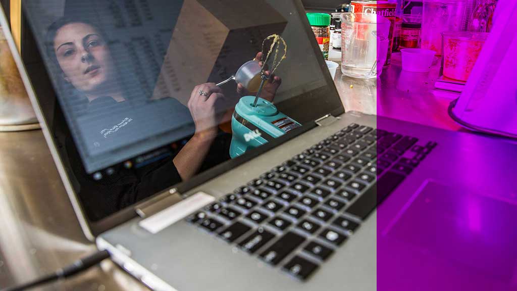 A chef looking at her laptop screen while holding a mixer 